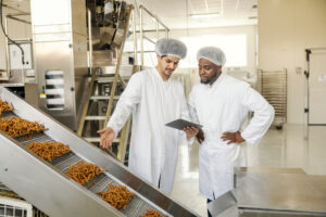 Interracial supervisor with tablet doing quality control check of food on food processing machine and talking with food factory worker.
